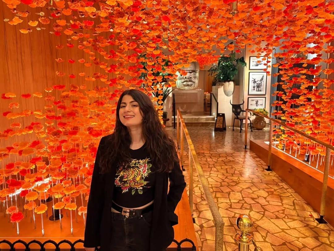 a photo of a dark haired woman in a black blazer standing in front of an ornate orange mezanine with a big smile on her face